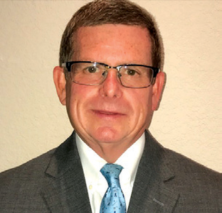 Man in a dark suit, light blue tie, and glasses, posing against a plain beige background