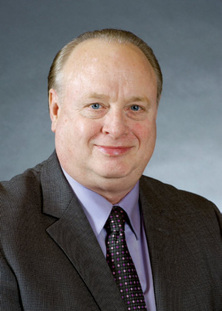 Studio headshot of a man in a dark suit, lavender shirt, and patterned tie, smiling against a gray background