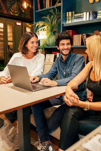 Three coworkers chatting at a table with a laptop in a cozy office lounge