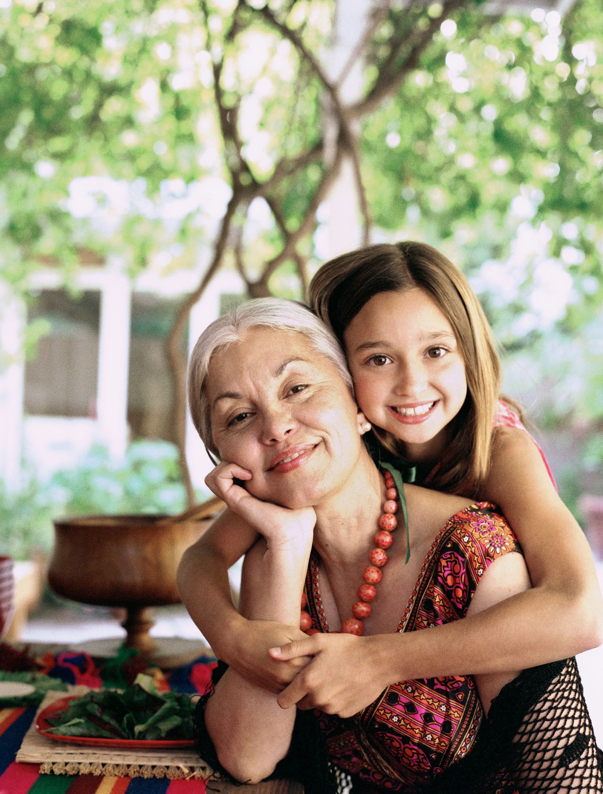 Smiling older woman and girl hugging outdoors under a tree, with colorful cushions nearby