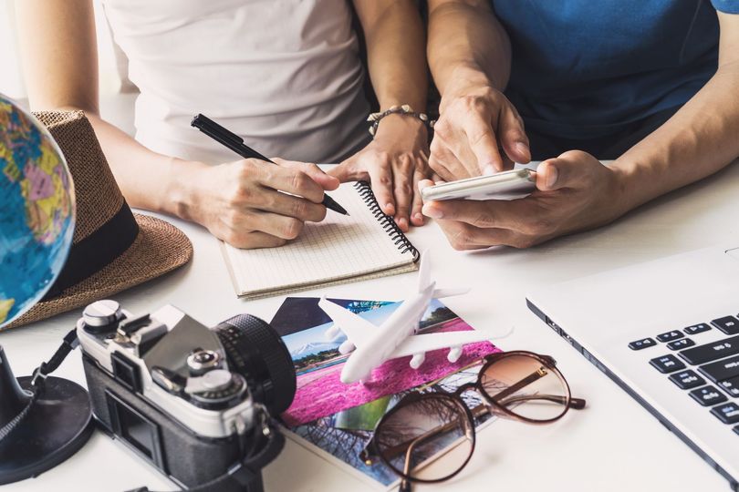 Hands using a notebook and smartphone at a desk with a camera, glasses, and laptop nearby
