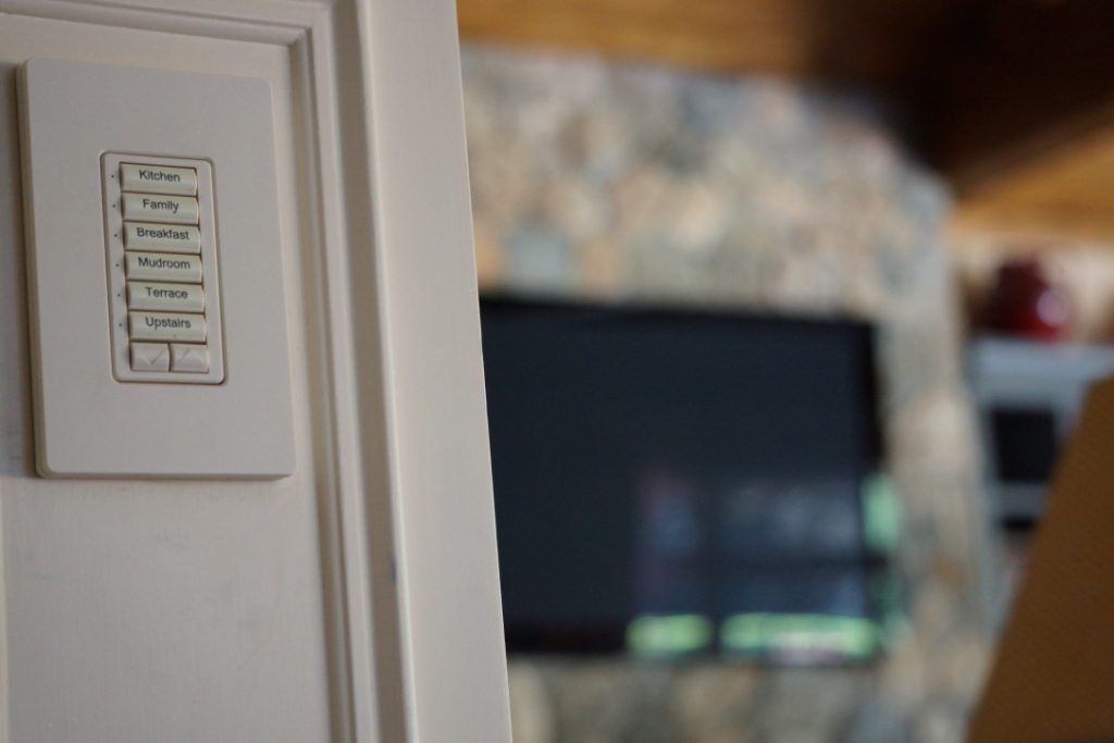 White light switch panel on a door; TV and stone fireplace blurred in background.