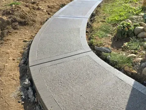 A curved concrete walkway featuring a textured center panel with smooth borders, situated between dirt and grass.