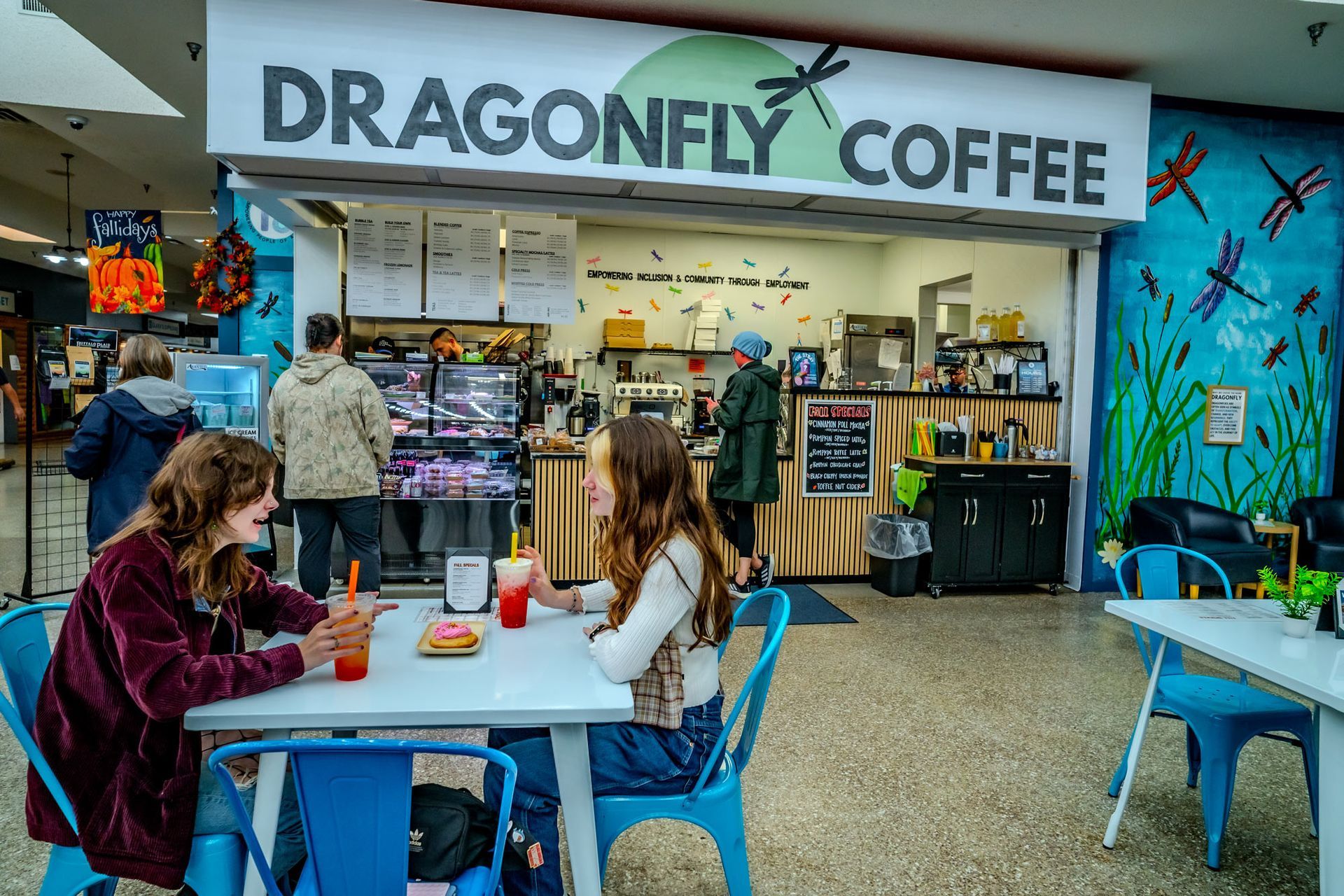 Two people at Dragonfly Coffee, seated at a table. Shop has dragonfly mural and menu boards.
