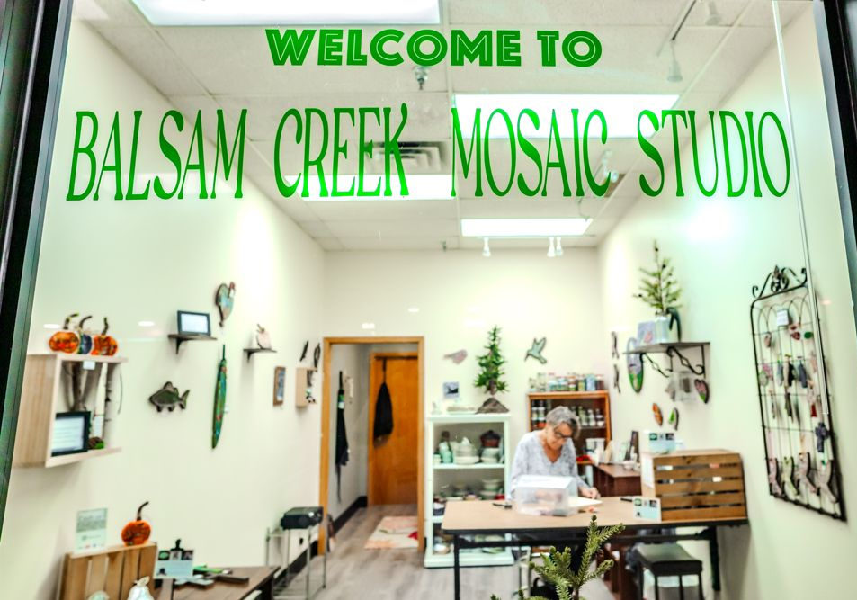 Window display of a mosaic studio: green lettering, woman working at a table, various mosaics on the wall.