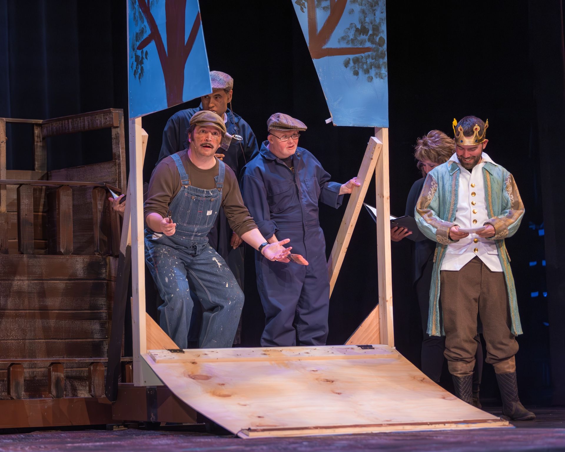 Actors on a stage, some in overalls, others in costume, a wooden set piece.