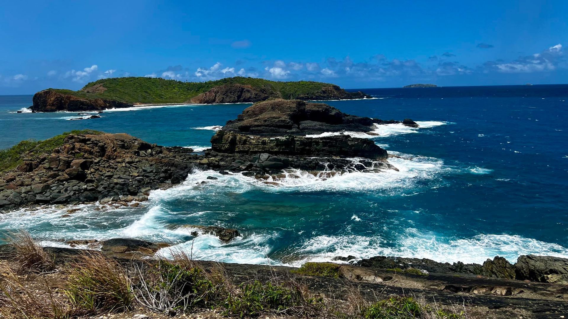 Rocky coastline with waves crashing against dark rocks, with islands in the distance under a blue sky.