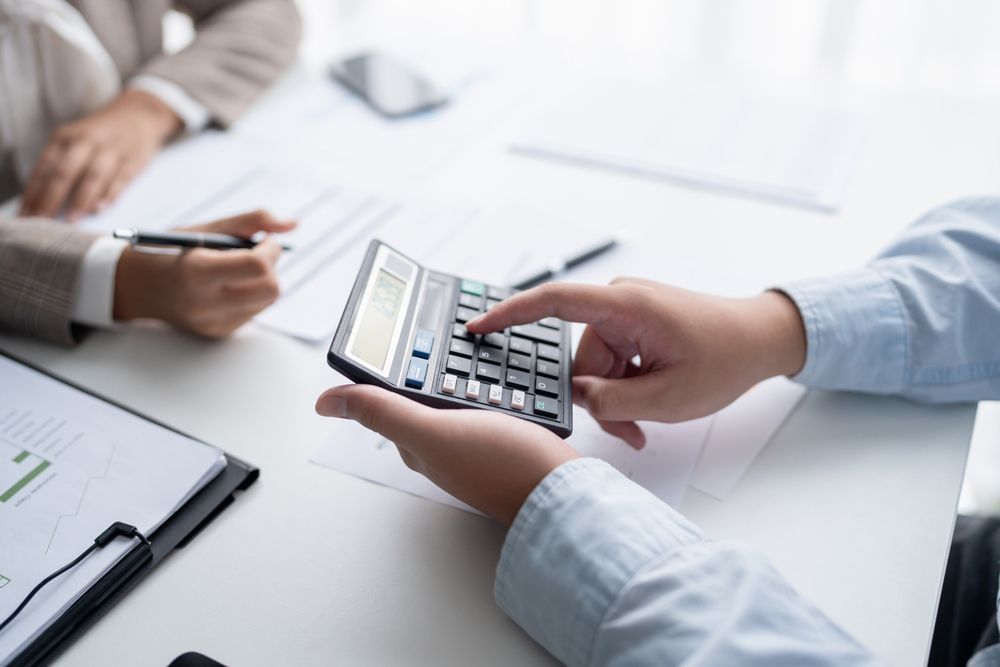 Person using a calculator at a desk, reviewing documents with another person.