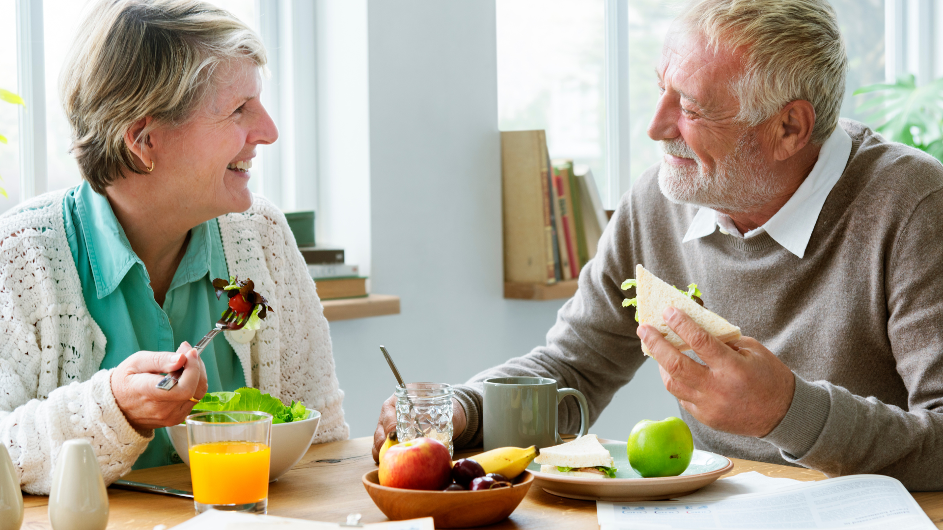 Two people sit at a table in a bright, sunlit room, smiling and talking while eating a healthy lunch together.