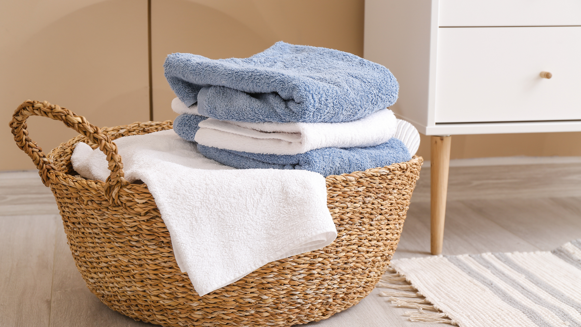A woven wicker laundry basket holds a stack of blue and white folded towels, sitting on a floor near a white cabinet.