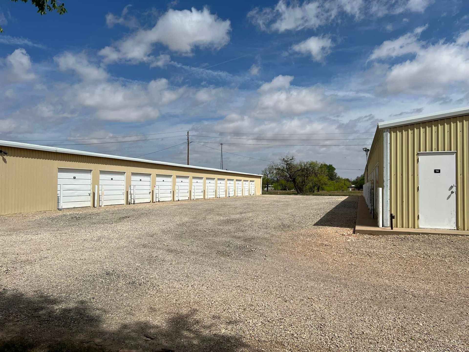 Storage units under a bright blue sky with scattered clouds. Gravel parking area.