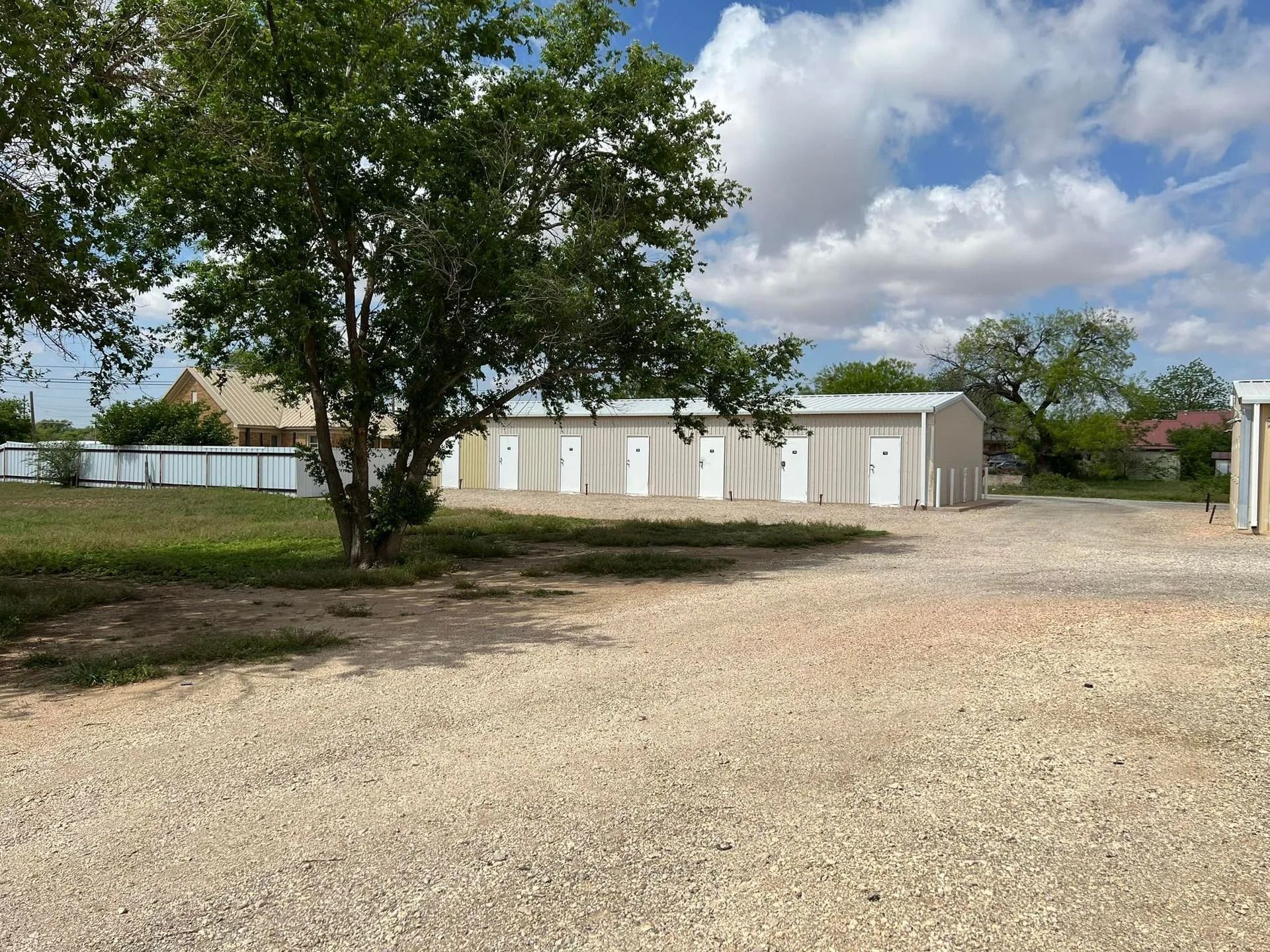 Storage units in a gravel lot under a partly cloudy sky. Tree in the foreground.