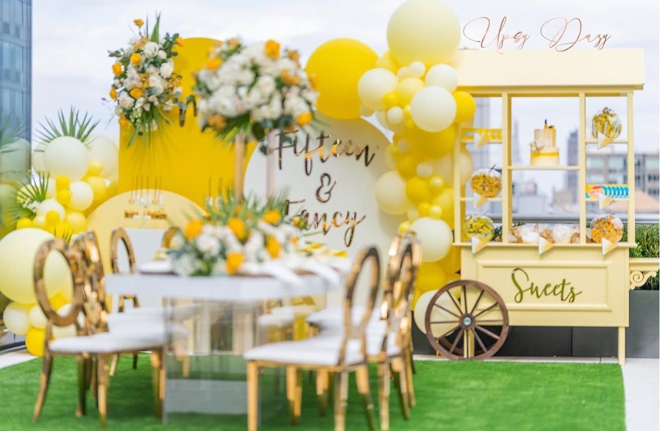 A table and chairs are decorated with yellow and white balloons and flowers.