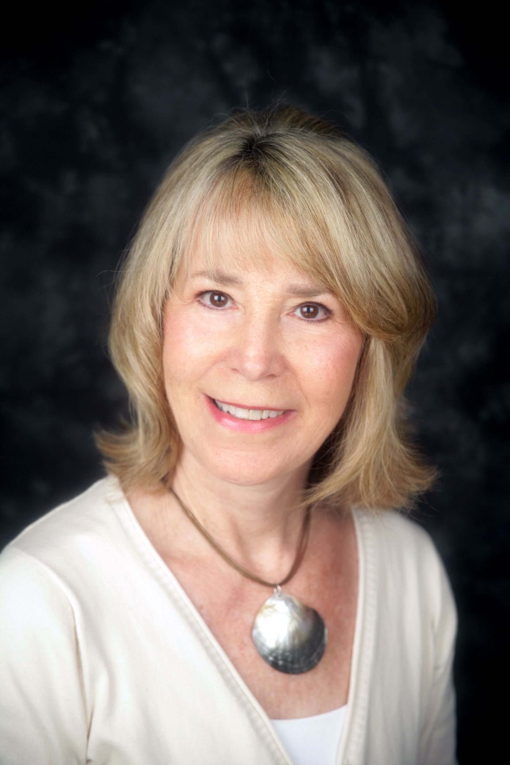 A person with shoulder-length blonde hair wearing a white top and a large, round metallic pendant necklace.