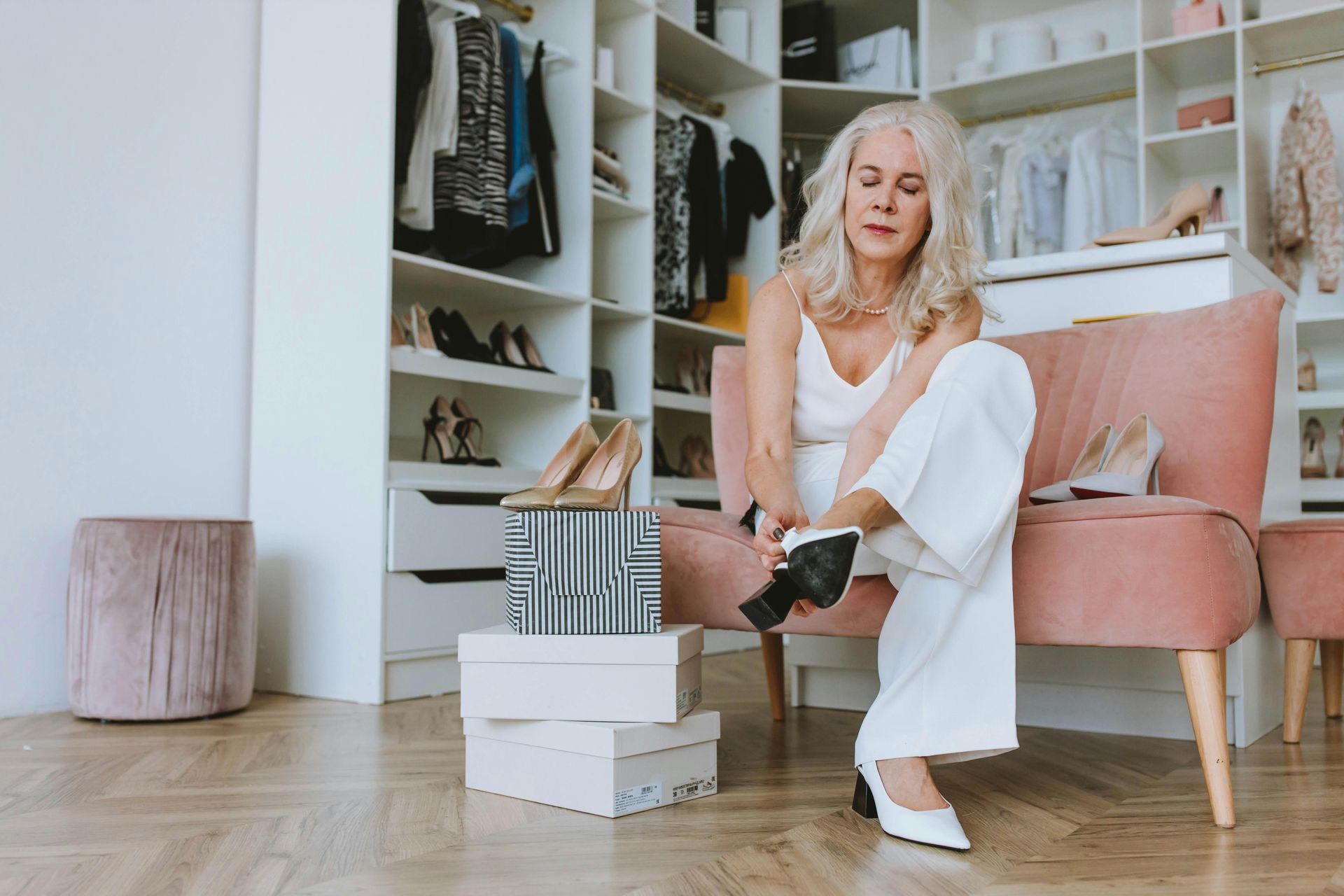 A person sitting on a pink velvet chair in a closet, trying on white high-heeled shoes next to stacked shoe boxes.