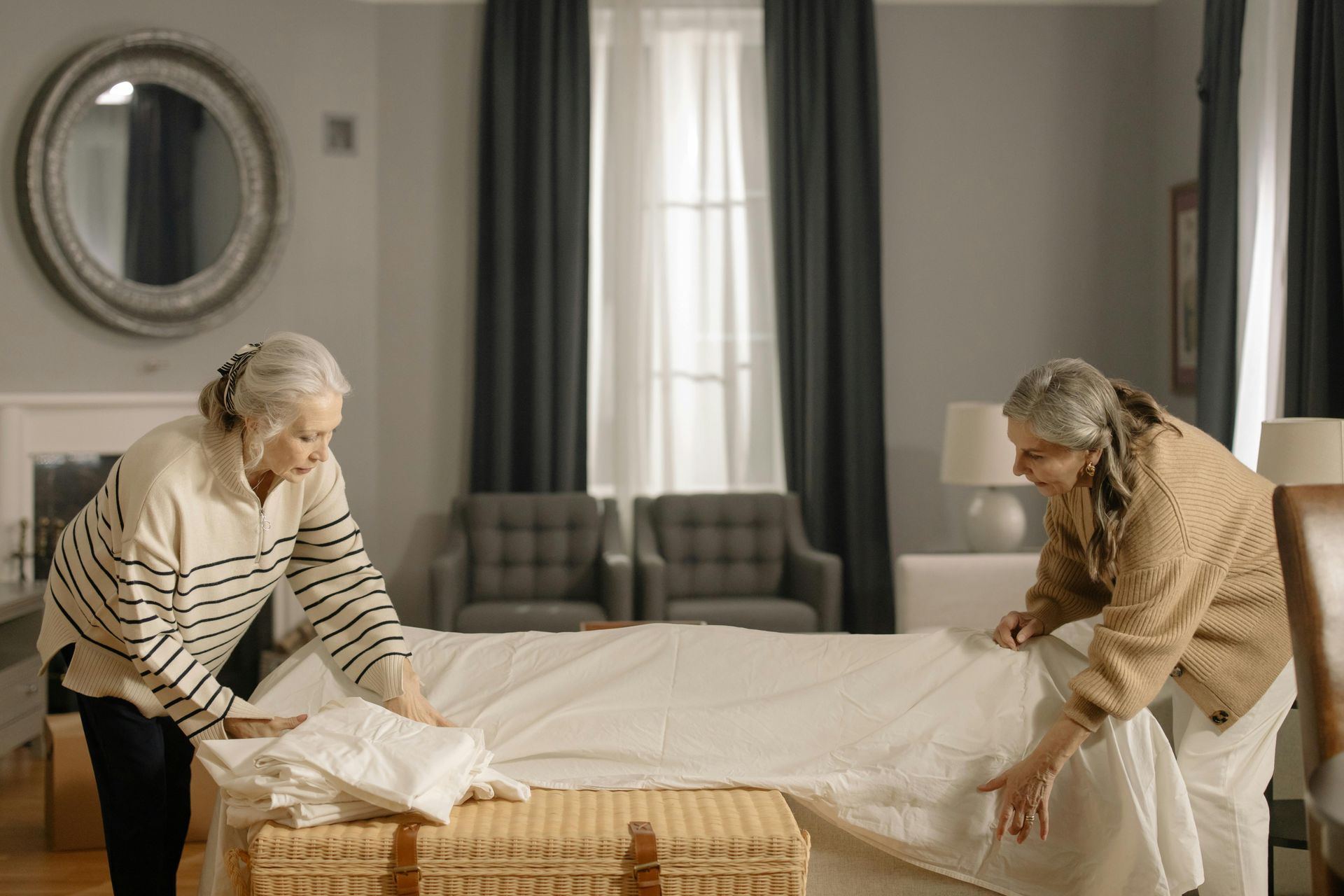 Two people stand on opposite sides of a bed, smoothing a white sheet over a woven trunk in a neutral-toned room.