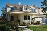 Two-story beige house with a porch, garage, and green lawn under a blue sky.