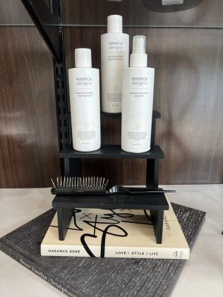 Three white hair care products on a black shelf, with a comb and book below.