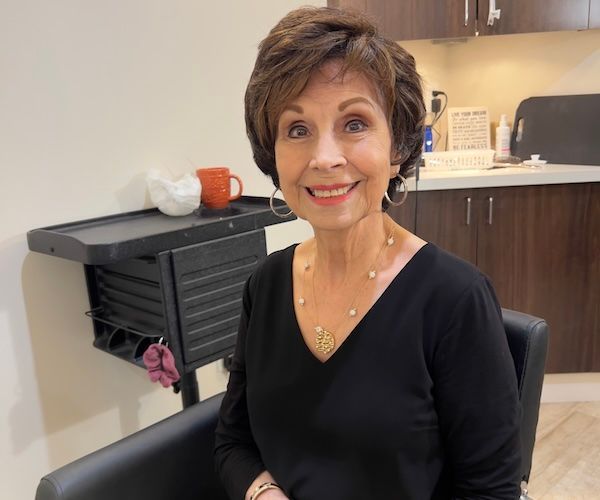 Woman with short brown hair smiles, wearing a black top, sitting in a chair in a salon.