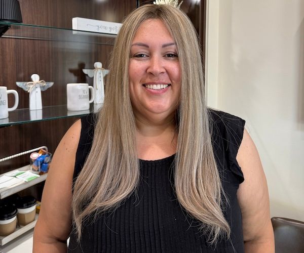 Woman with blonde hair smiling in a salon. She wears a black top. Background includes shelves with decor.