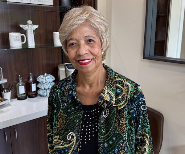 Woman in patterned jacket smiles, standing near a shelf with decorative items.