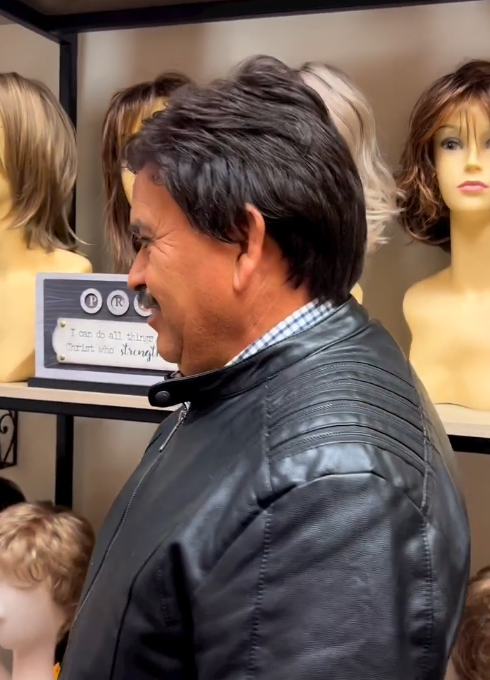 Man in black jacket, looking right, in a wig shop with multiple wigs on mannequins in the background.