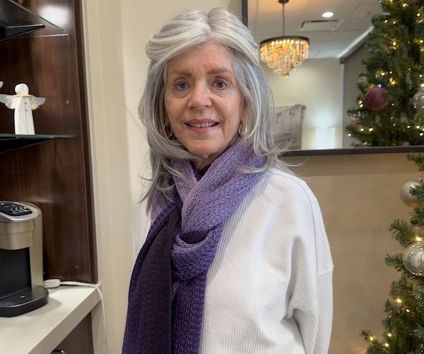 Woman with gray hair wearing a purple scarf and white sweater, standing near a Christmas tree.