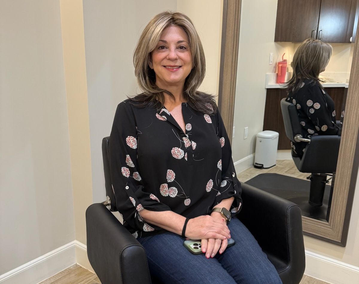 Woman in a salon chair smiles, wearing a black floral top and jeans. She sits in front of a mirror.