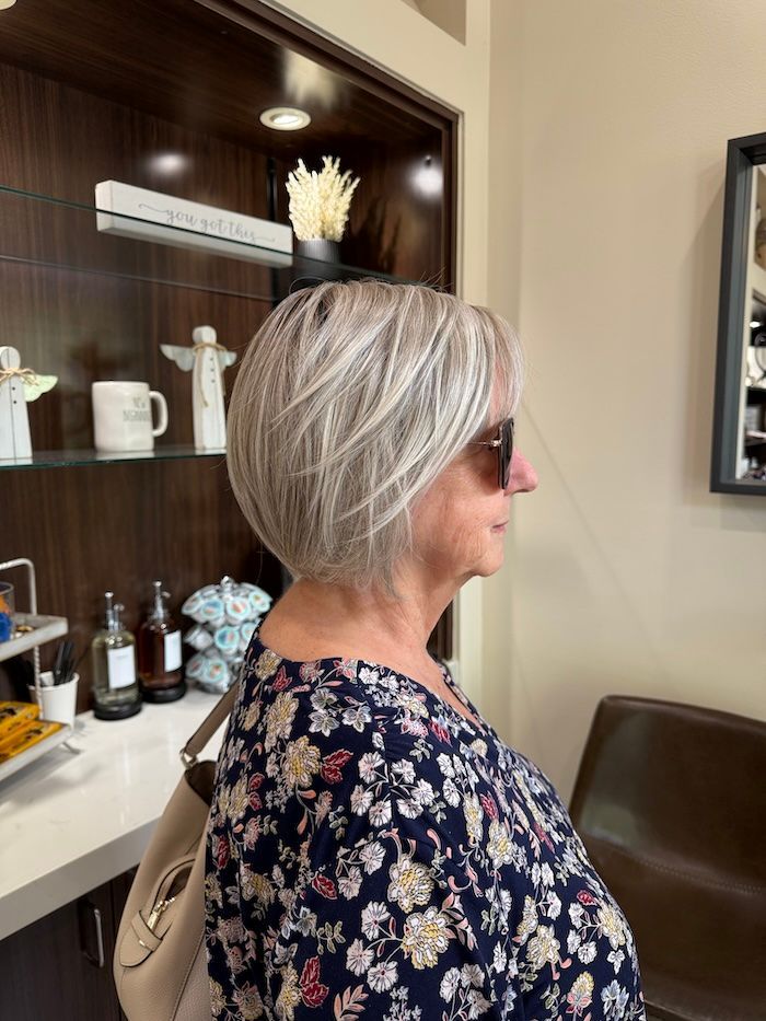 Woman with short, layered gray hair wearing sunglasses, stands in front of a shelf in a salon.