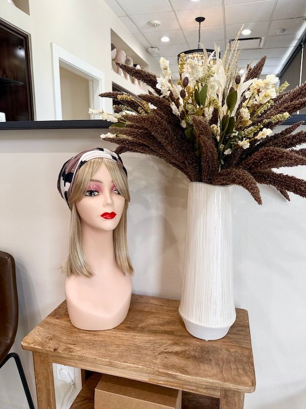 Mannequin head with blonde wig and patterned headband sits on a wooden table next to a white vase of dried flowers.