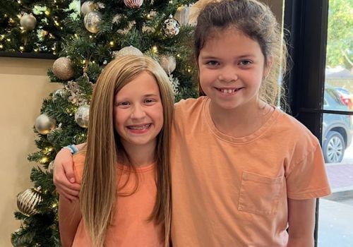 Two girls smiling, posing by a decorated Christmas tree. One has long blonde hair, the other's is dark and pulled back.