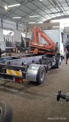 Un escavatore arancione su telaio di camion all'interno di un magazzino con travi metalliche e attrezzature.