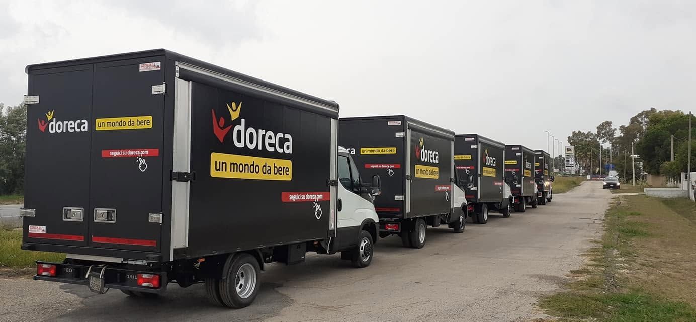 Camion neri per le consegne allineati sul ciglio della strada, con una banchina sterrata e degli alberi sullo sfondo.