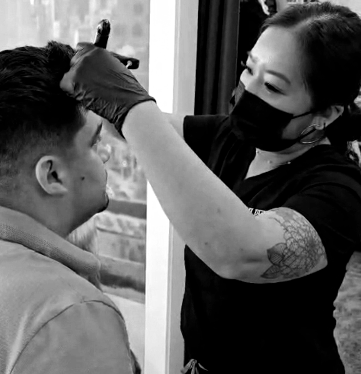 Masked stylist trims a man's hair near a window in black-and-white