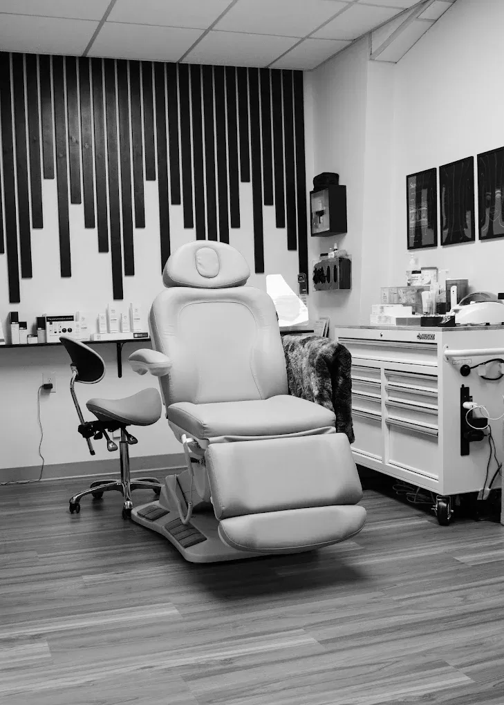 A treatment room featuring a beige reclining chair, a rolling stool, and a white storage cart on a wood floor.
