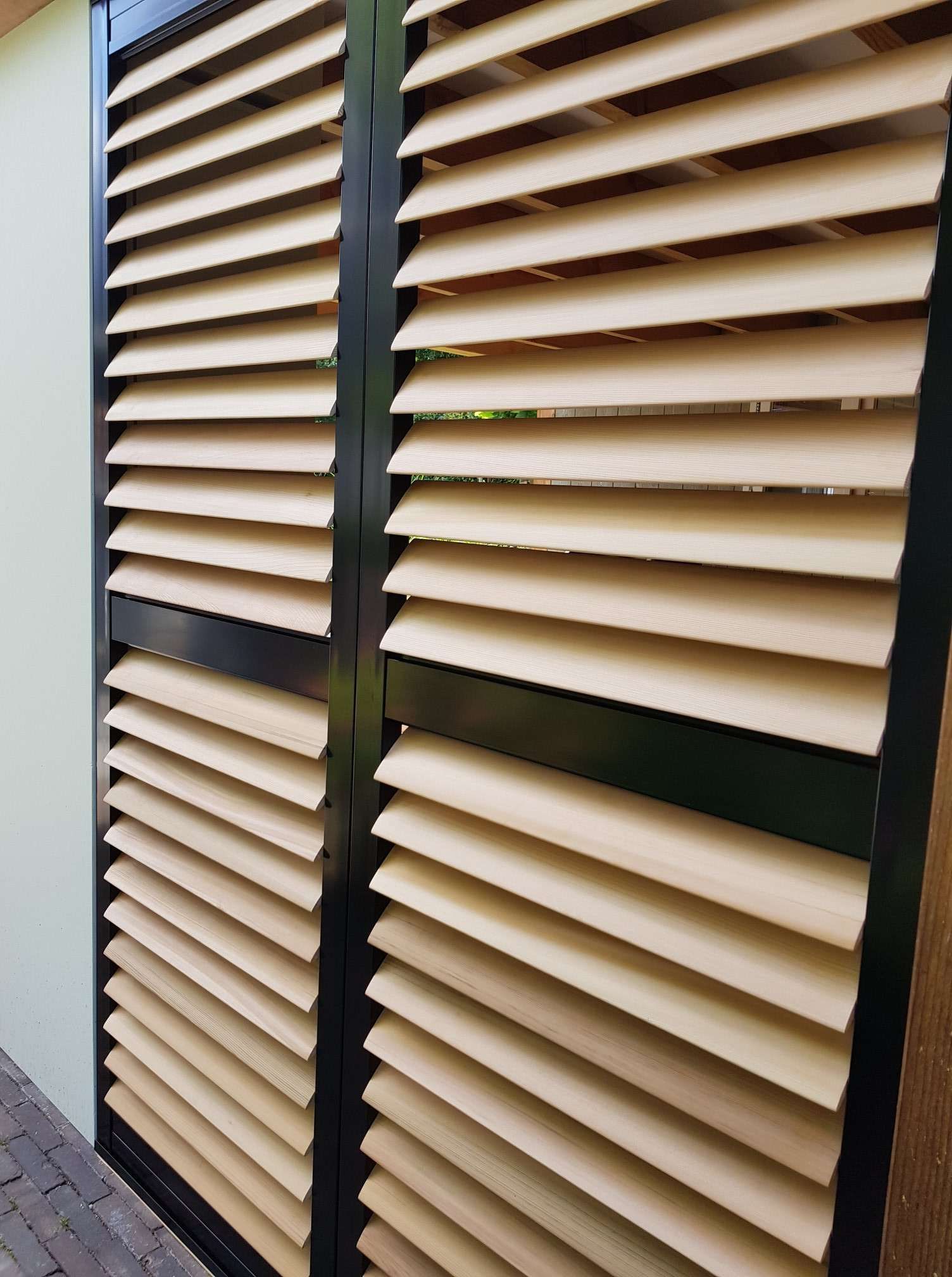 Beige louvered shutters in dark green frames covering a window on a brick exterior wall — Fineworx Joinery in Warnervale, NSW