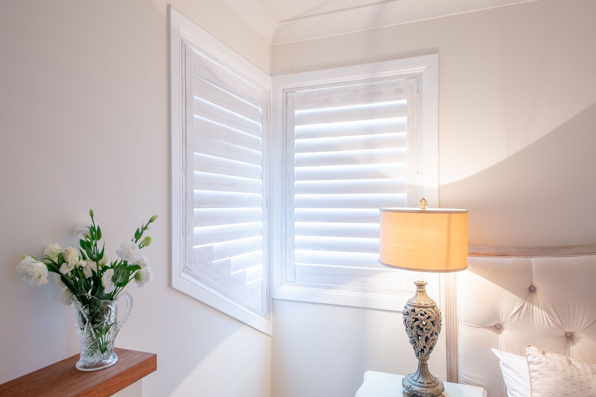Bright bedroom corner with white shutters, a lit table lamp, tufted headboard, and flowers on a dresser — Fineworx Joinery in Warnervale, NSW