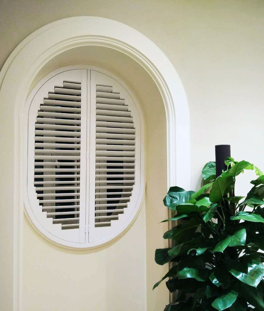 Arched window with white shutters beside a green potted plant on a cream wall — Fineworx Joinery in Warnervale, NSW