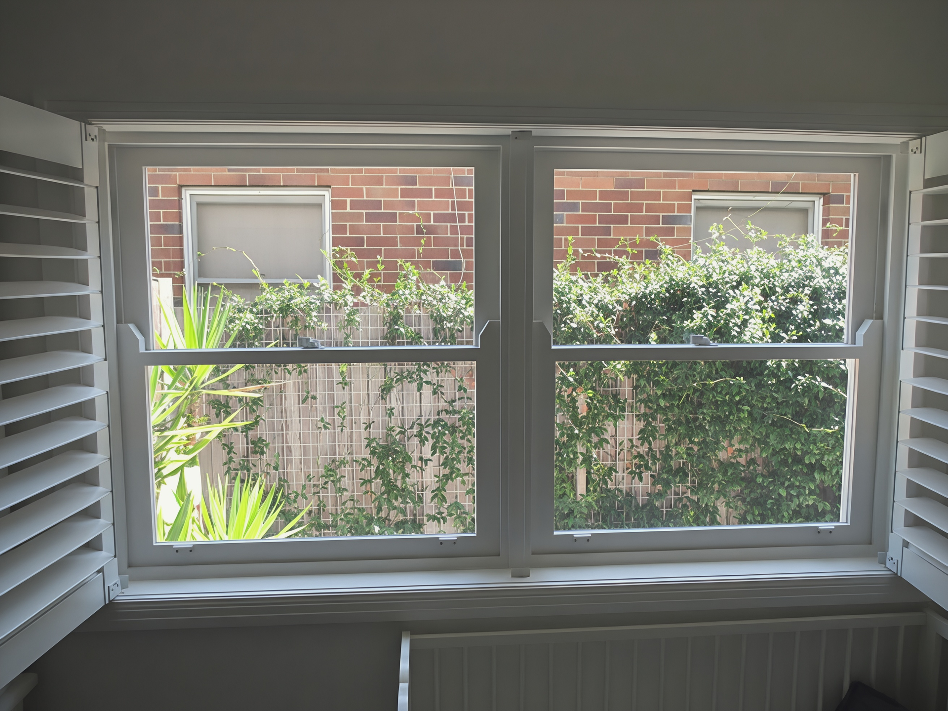 Window view of a brick building, shrubs, and a small fenced garden, framed by white shutters and blinds — Fineworx Joinery in Warnervale, NSW