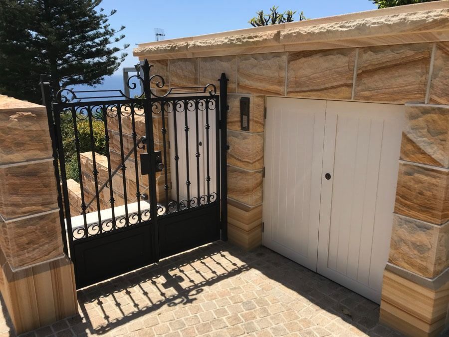 Black Wrought Iron Gate Beside a White Garage Door in A Sunlit Stone Wall — Fineworx Joinery in Warnervale, NSW
