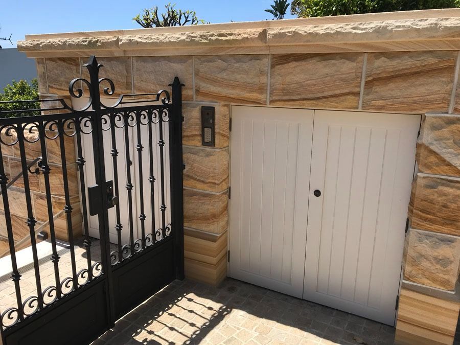 White Garage Door Beside Black Wrought Iron Gate and Stone Wall in A Sunny Driveway — Fineworx Joinery in Warnervale, NSW