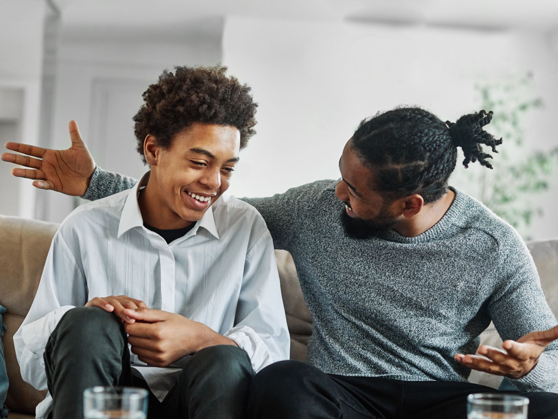 Two people on a couch, one laughing with an arm around him, the other gesturing with a smile.