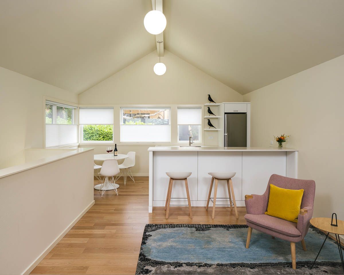 Bright modern open-plan living room with white walls, wood floors, bar stools, and a pink chair on a blue rug