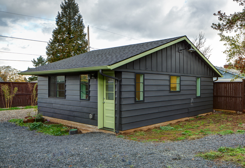 Small dark gray house with a light green door and trim, shown on a gravel lot under cloudy skies