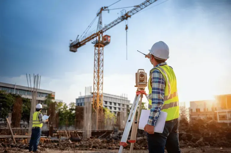 Construction workers in safety vests and hard hats at a building site with a crane and surveying tripod