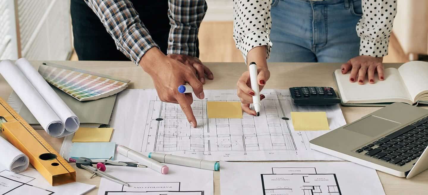 Two people look at plans on a desk. They're holding markers and pointing at things.