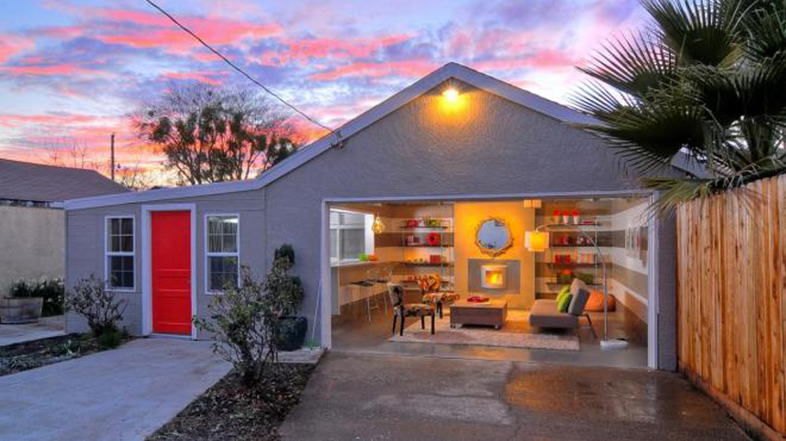 Gray house at sunset with red front door, lit garage/workshop, and wooden fence in front yard