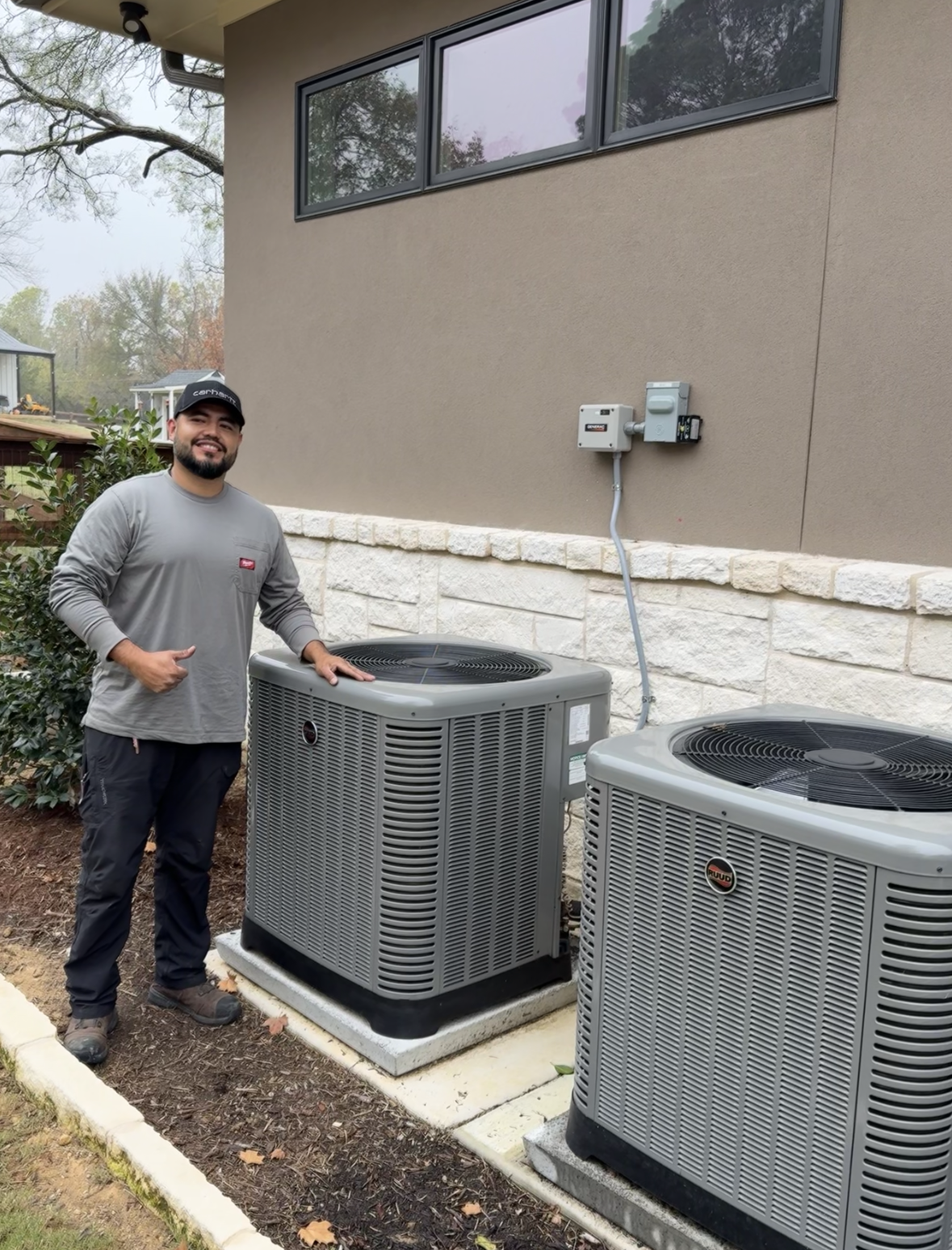 Man standing next to two gray HVAC units, pointing at them, exterior of a modern building.