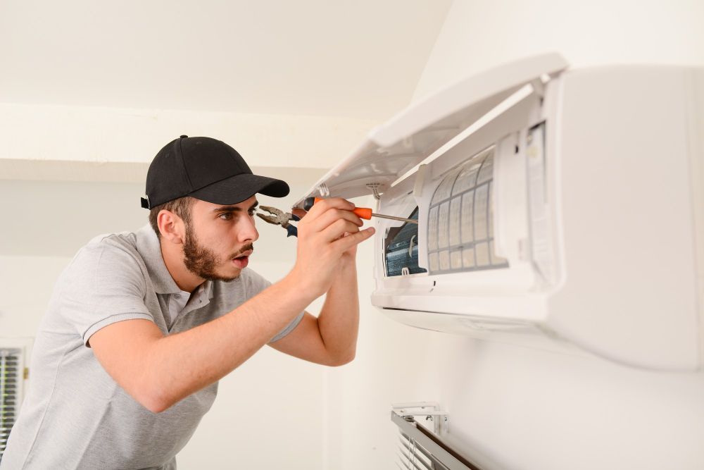 Man in cap repairing an air conditioner on a white wall, using tools.