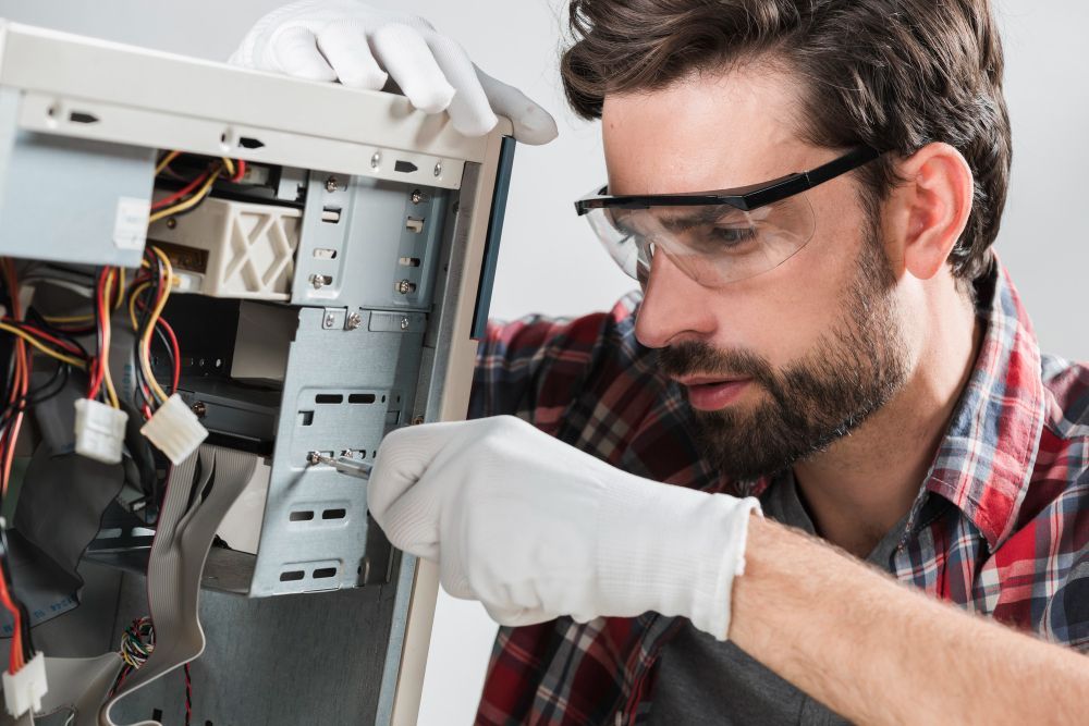 Man fixing a desktop computer, wearing safety glasses and gloves, in a well-lit room.